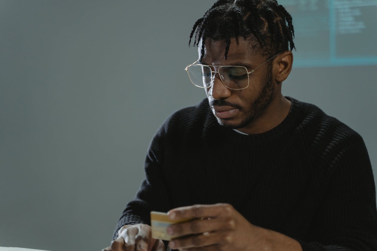Focused man with eyeglasses holding a credit card in a dimly lit room, wearing a black sweater.
